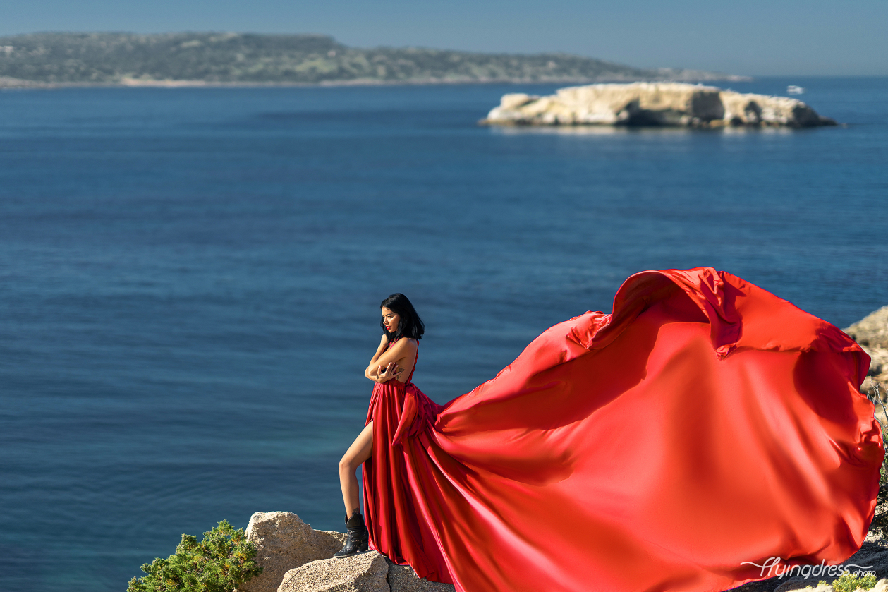 A woman in a flowing red dress and black boots stands elegantly on rocky cliffs with the expansive blue sea and distant islands of Kavouri, Athens in the background, creating a dramatic and captivating scene.